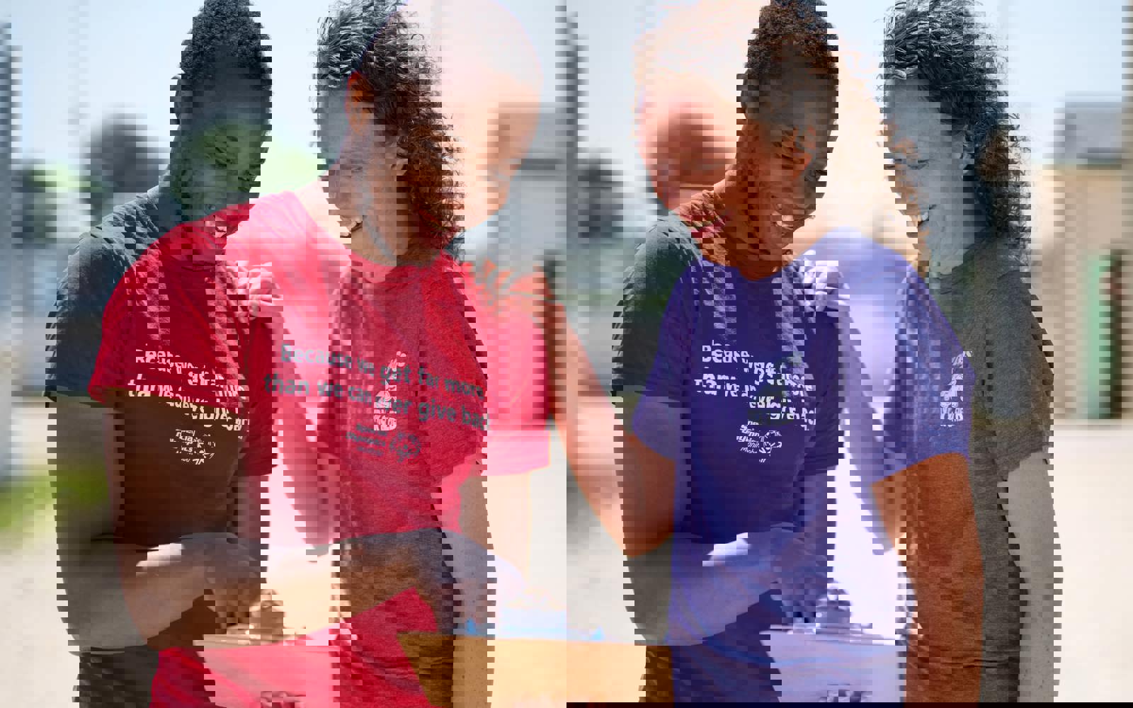 A person in a red shirt with the words "Because we get far more than we can ever give back" holding and pointing at a clipboard while a person in a purple shirt smiles down at the clipboard. 